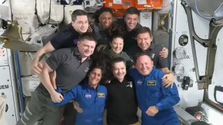 nine astronauts post for a picture while floating in zero gravity in a cramped room filed with computer screens and white boxes
