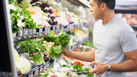 Man in a grocery store inspecting leafy greens and holding a shopping basket
