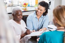 African American older female patient signs medical forms during home healthcare visit. The woman's adult daughter is helping her with the form. A home healthcare nurse is in the foreground.