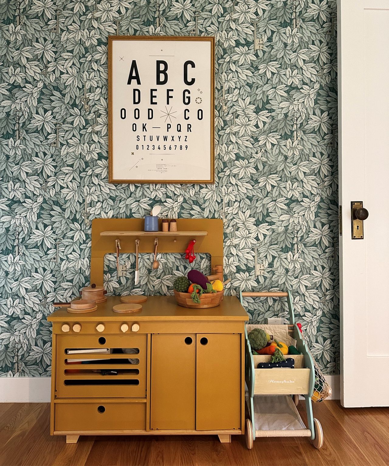 A yellow children's play kitchen in a room with botanical wallpaper and wooden floors