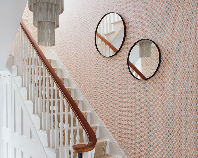 Staircase with wallpapered wall, orange patterned wallpaper, two round mirrors and low hanging chandelier