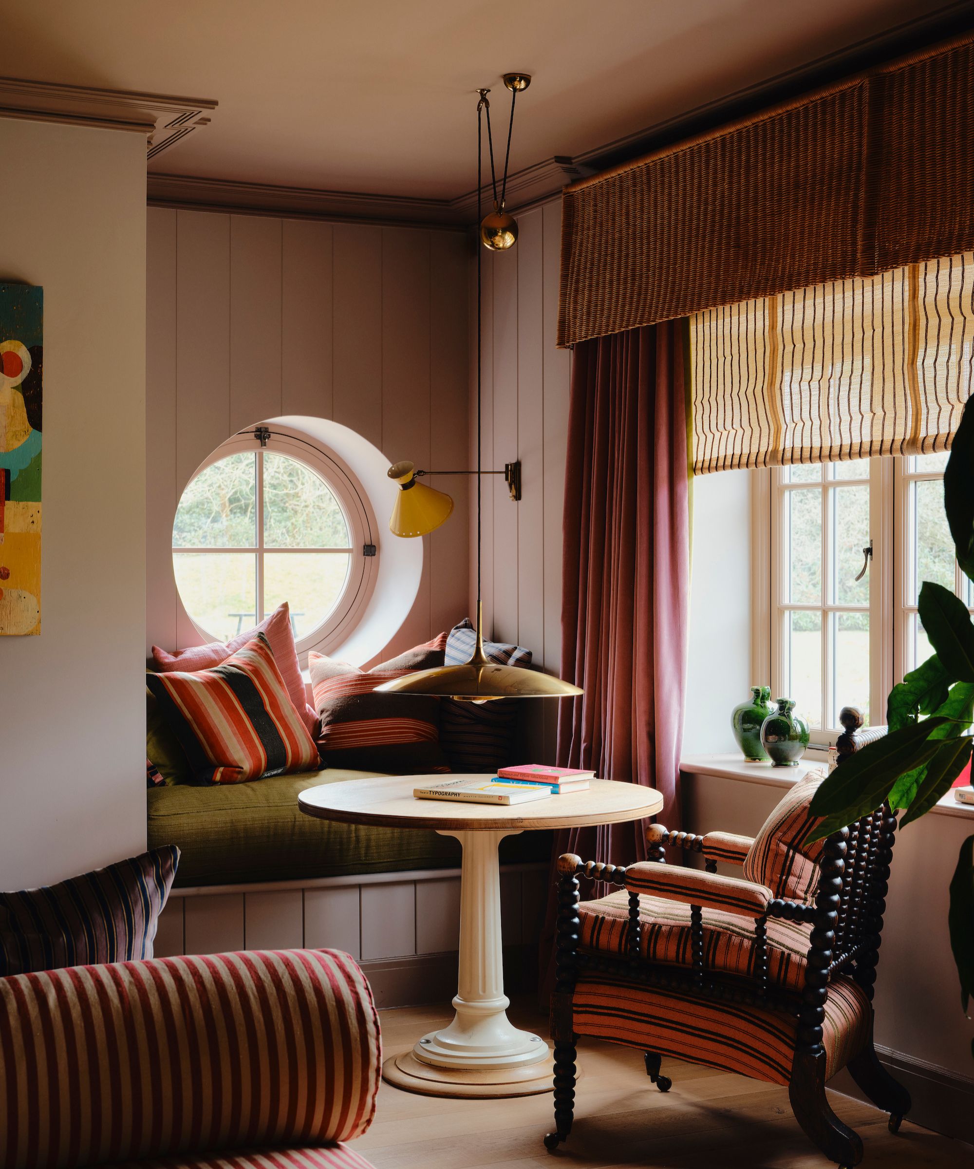 Reading nook with pale pink painted shiplap walls, round window, built-in bench with green cushion, round wooden table, bobbin armchair and a large window with a rattan blind and purple drapes