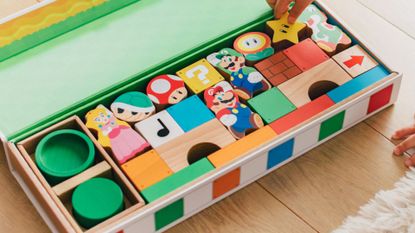 A hand reaching to take a wooden Star block from a tray filled with wooden Super Mario characters and items