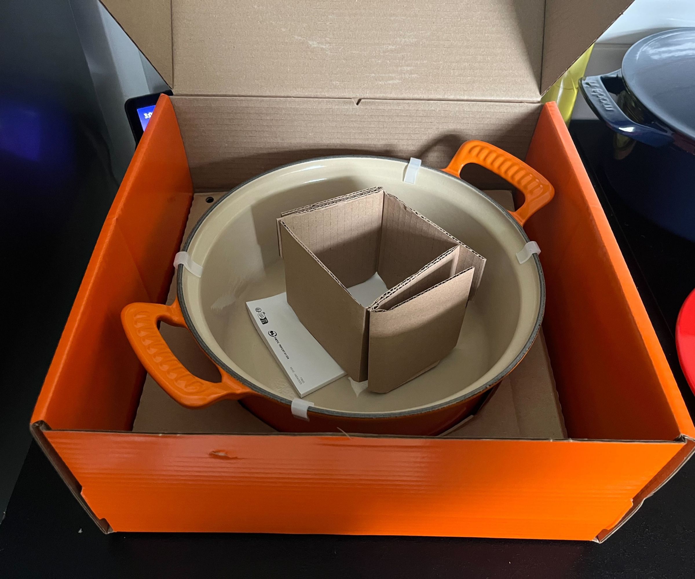An open orange Le Creuset box with an orange Dutch oven inside, with a square of cardboard in the centre