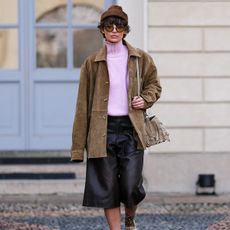 A woman wears a brown hat, suede jacket, pink turtleneck sweater, black bermuda shorts, and ivory moccasins. 