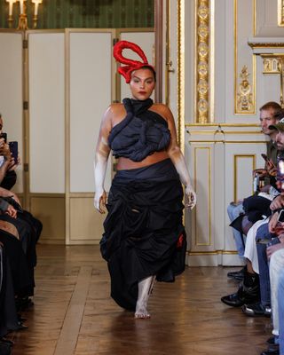 Paloma Elsesser walking in Torishéju Dumi Spring 2024 runway show wearing a sculptural black top, black skirt, a red head piece, and white hand and foot paint