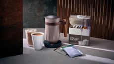 The new Hotel Chocolat velvetiser next to a cup of iced chocolate and hot chocolate, and flavor sachets