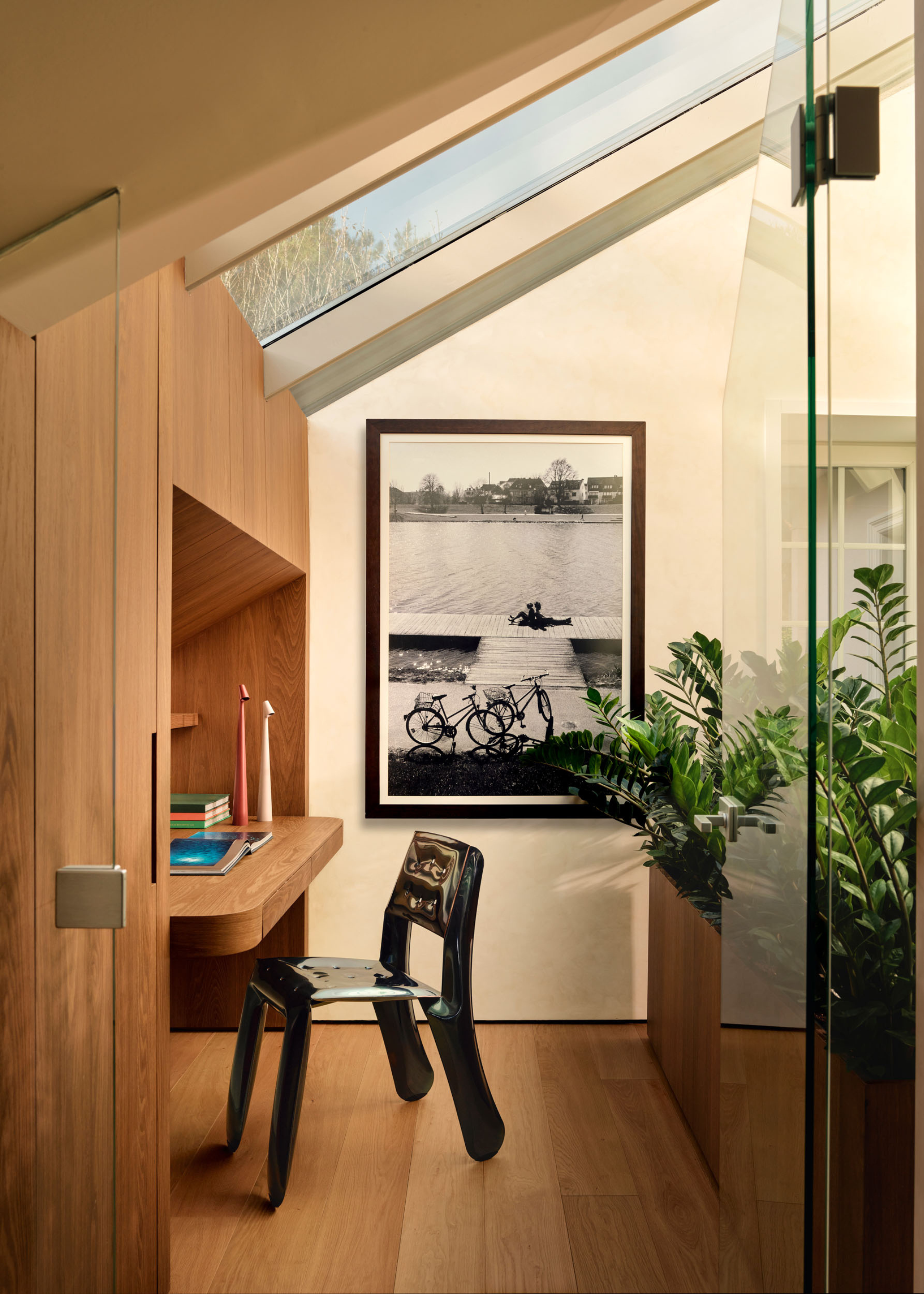 A study with a skylight, a wood floating desk and cupboards, a black and white print hanging on the wall, a planter with jade plants, and a black glossy metal chair