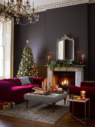 plum Christmas living room with magenta velvet couches, grey button backed footstool/coffee table, festive garland, mirror, candles, tree, chandelier