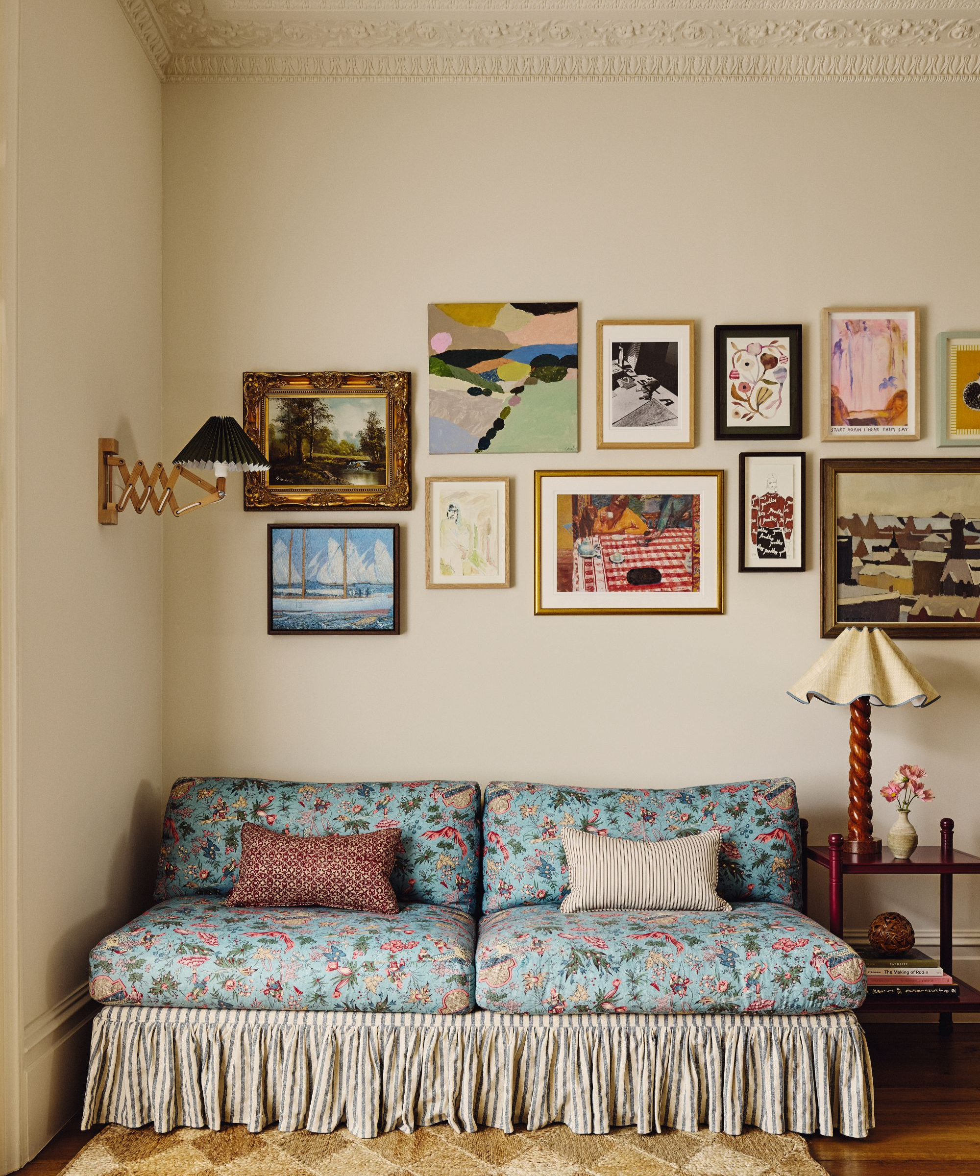 a sitting area with a blue floral printed sofa, a sofa skirt, two pillows, a gallery wall, a wall sconce, and a table lamp