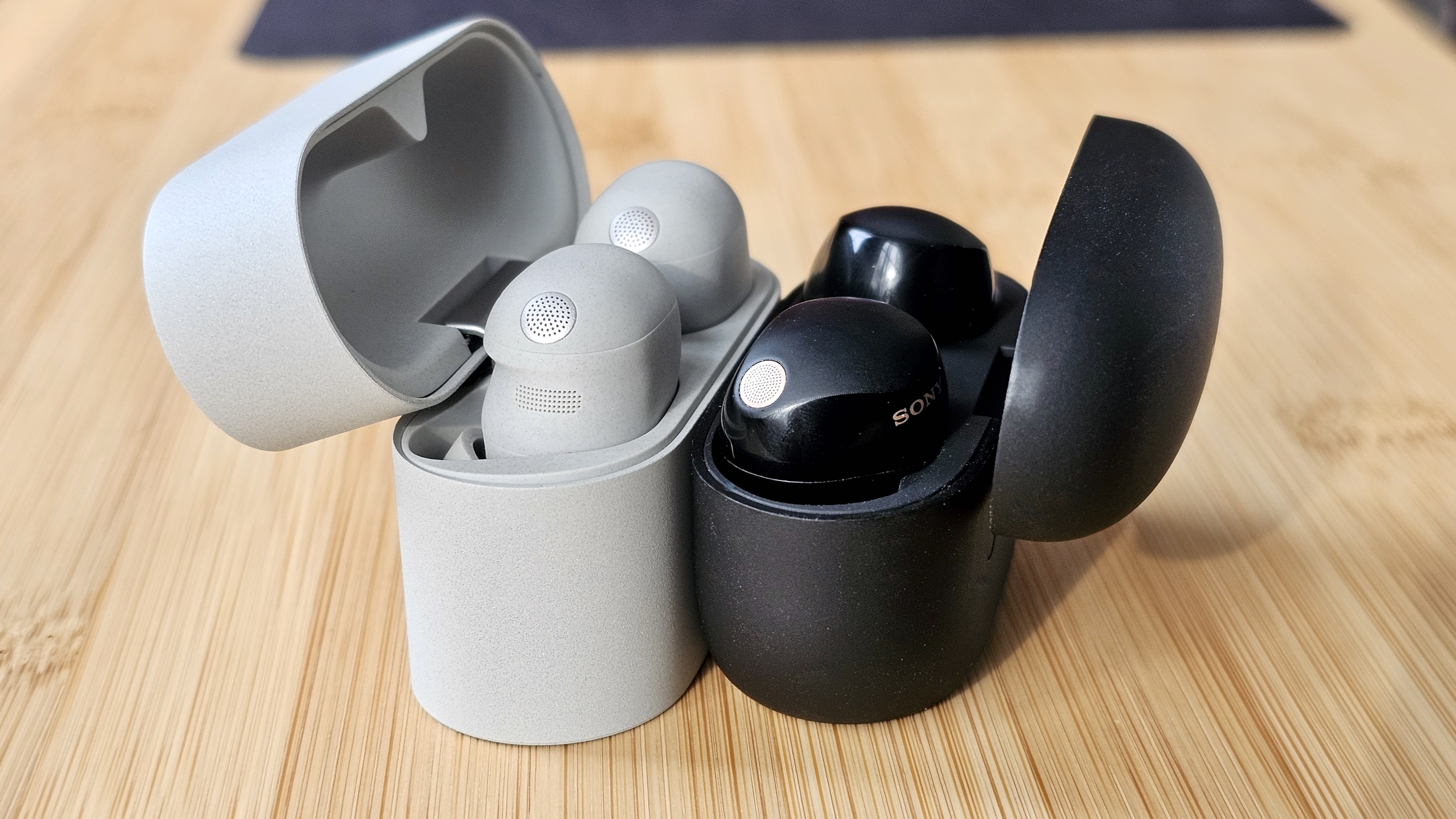 The Sony WF-1000XM6 and WF-1000XM5 side by side in a comparison, on a light wood table 