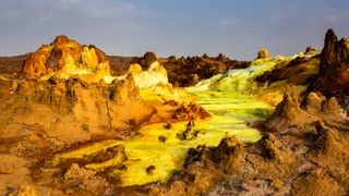 Strange landscape with brown and orange rocks surrounded by pools of yellowy green liquid.