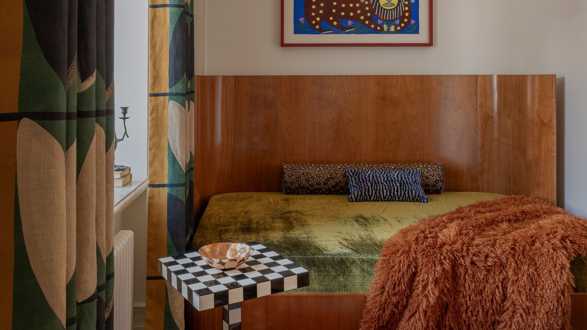 A pretty bedroom with a curved wood headboard, a velvetty bedscape with a bolster pillow, a furry throw, and a chequerboard side table