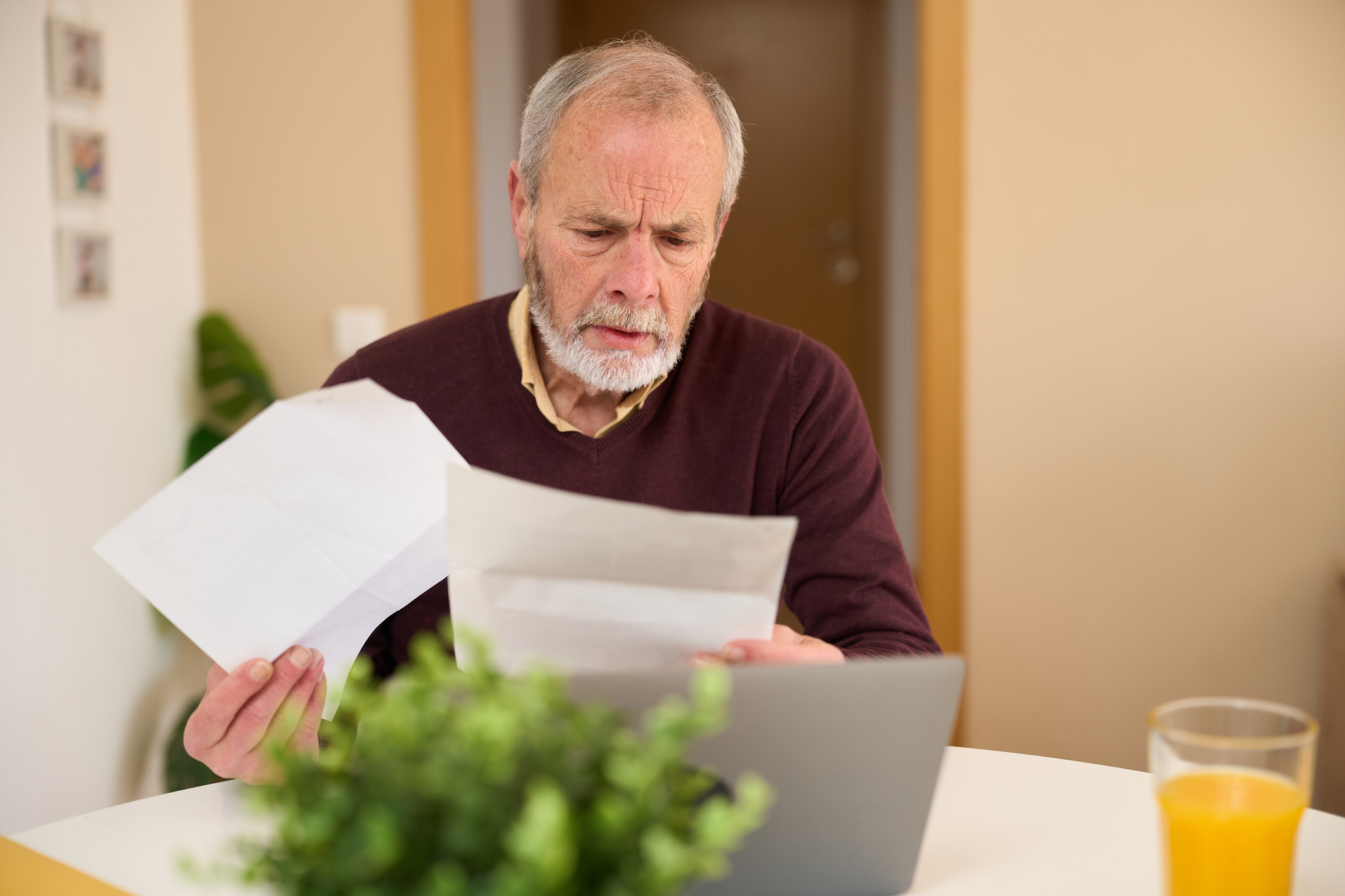 A senior man looks concerned while reviewing paperwork at his kitchen table.