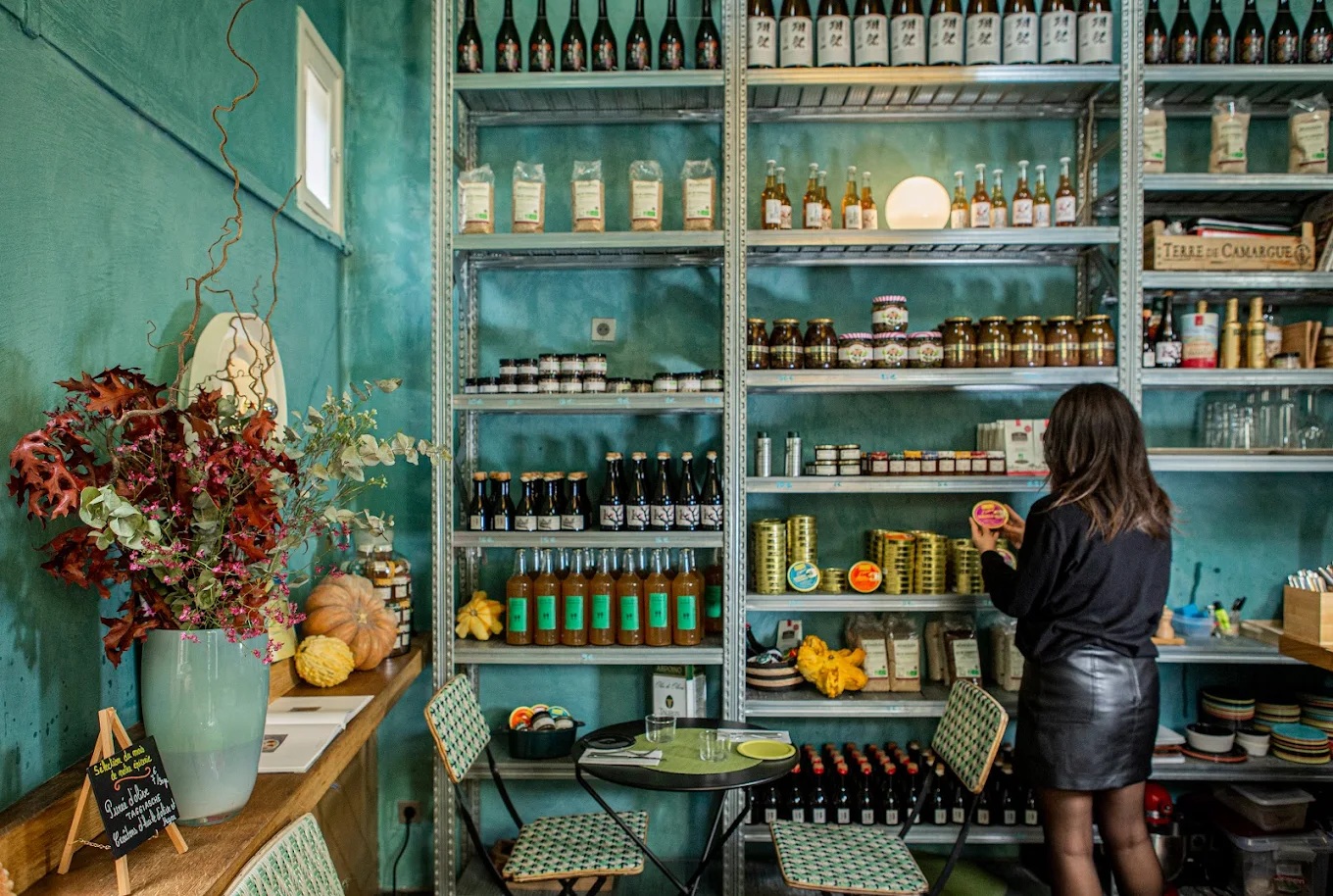 A modern deli boasts a tile-hued atmosphere, with metallic shelves stacked up with products which a woman is currently browsing.