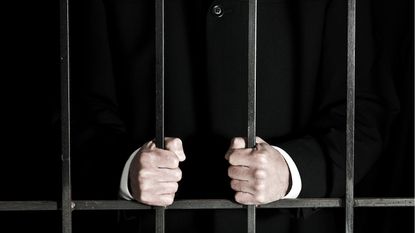 A business man in a jail cell, only his hands holding the bars showing.