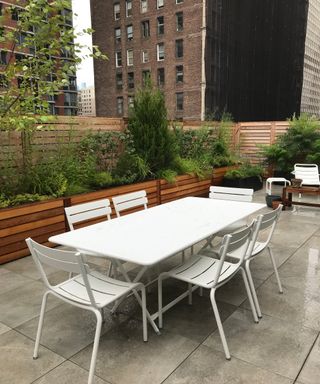 White metal outdoor dining table and 6 matching chairs in a narrow paver patio with planters around the edge
