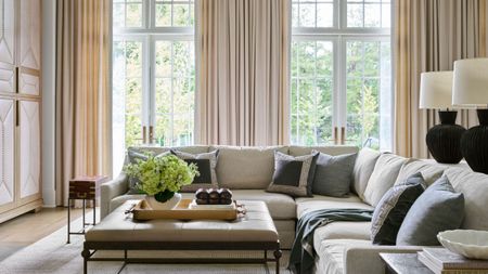 Modern living room with high ceilings and glass doors leading into the garden, long cream drapes, a large cream sectional sofa, grey pillows, a leather and a metal coffee table with decor on top.