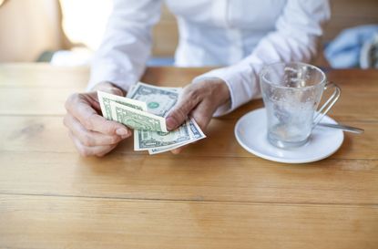 woman with white shirt ready to pay empty finished cappuccino coffee cup with dollar banknotes in hand on light brown wooden table cafe