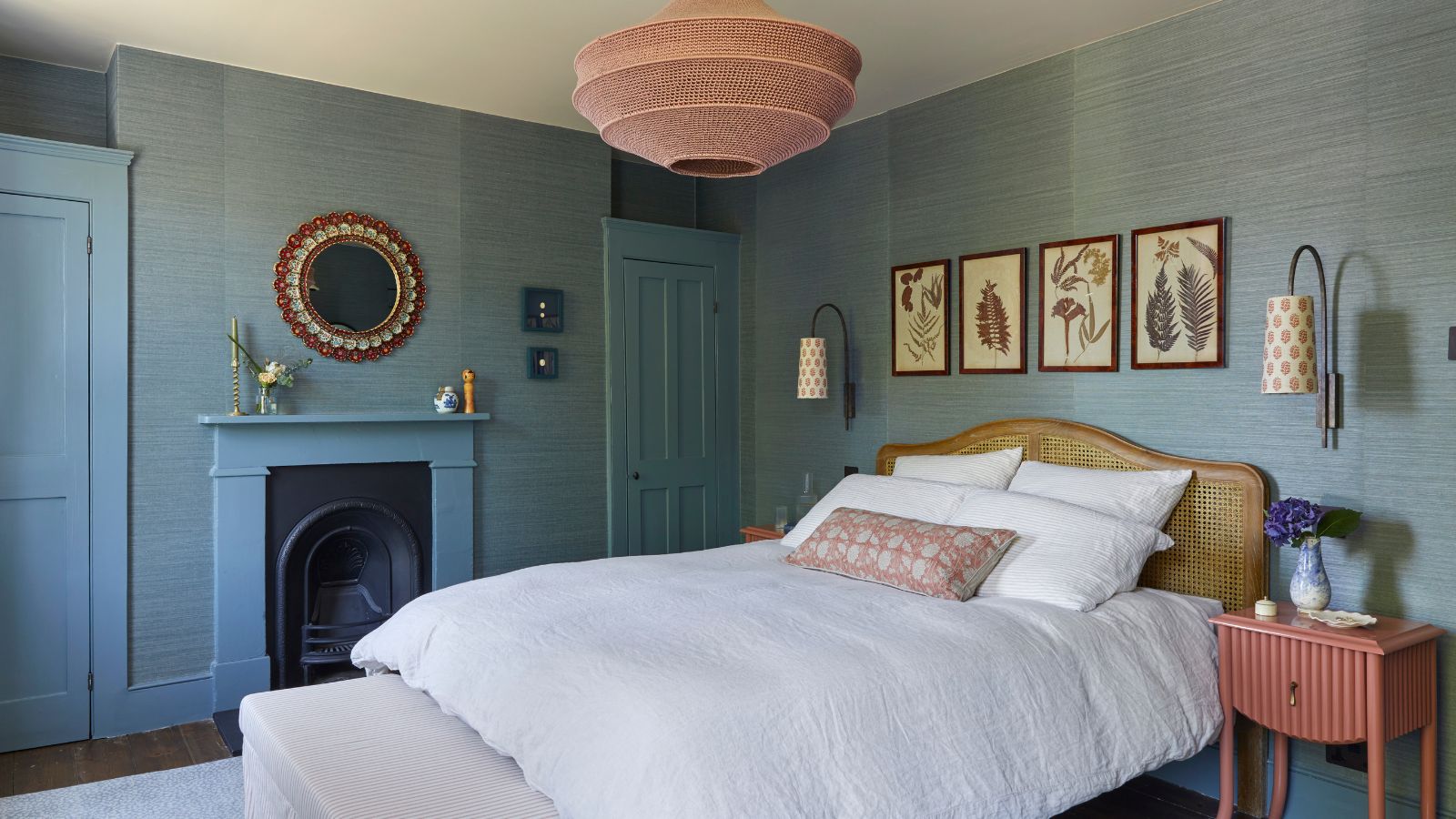 Bedroom with blue walls, blue painted fireplace, pink bedside table, rattan headboard, botanical prints and pink overhead light