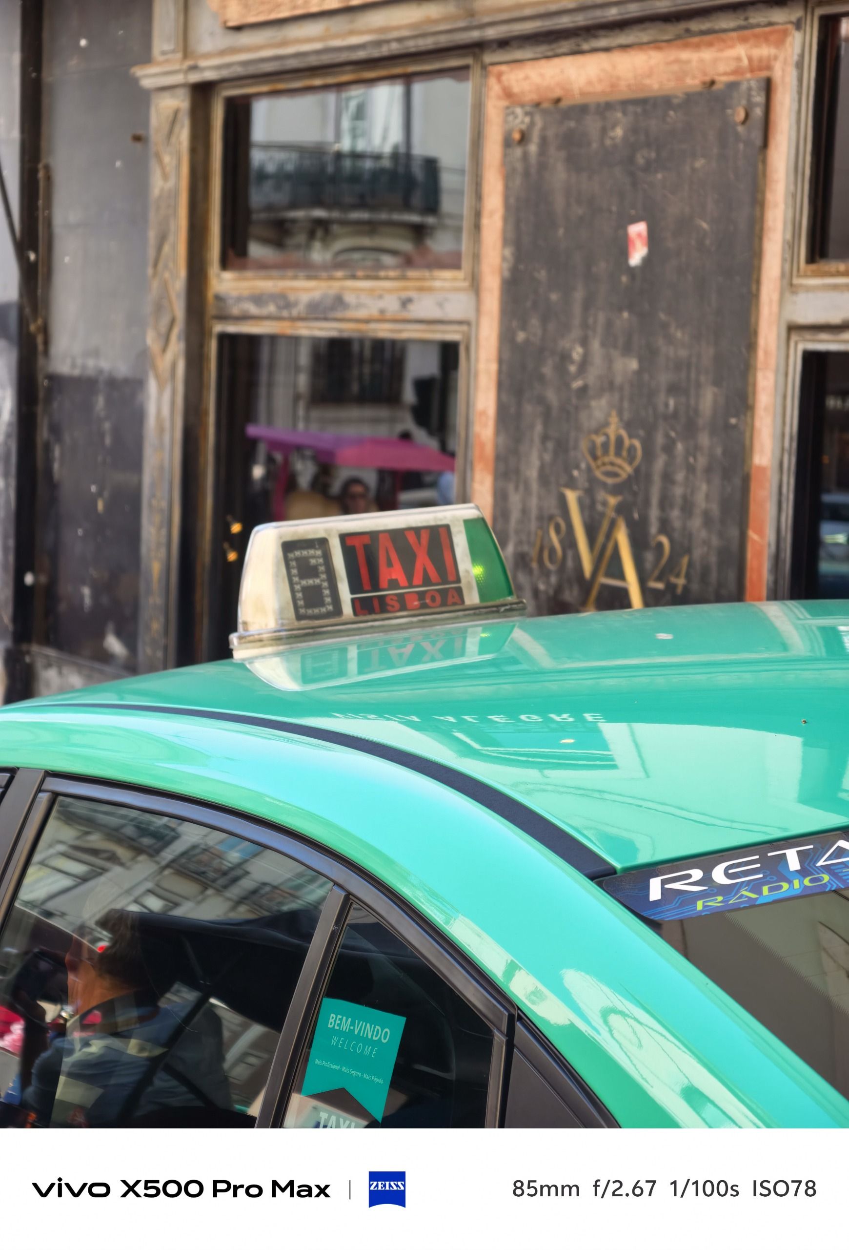 Close view of the roof sign on a turquoise Lisbon taxi, with an ornate building facade behind.