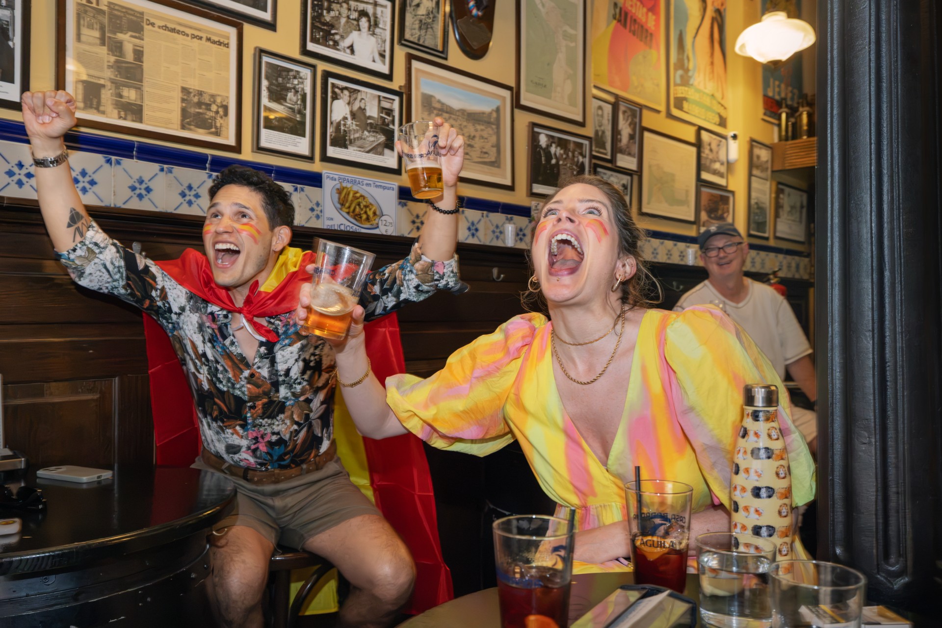 In a traditional Madrid bar, a man in a Spanish flag cape and a woman in a yellow and pink dress, both with red and yellow face paint, cheer ecstatically with beers raised as an older man smiles