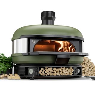 An original Gozney Dome pizza oven in olive green, surrounded with splits of wood and basil