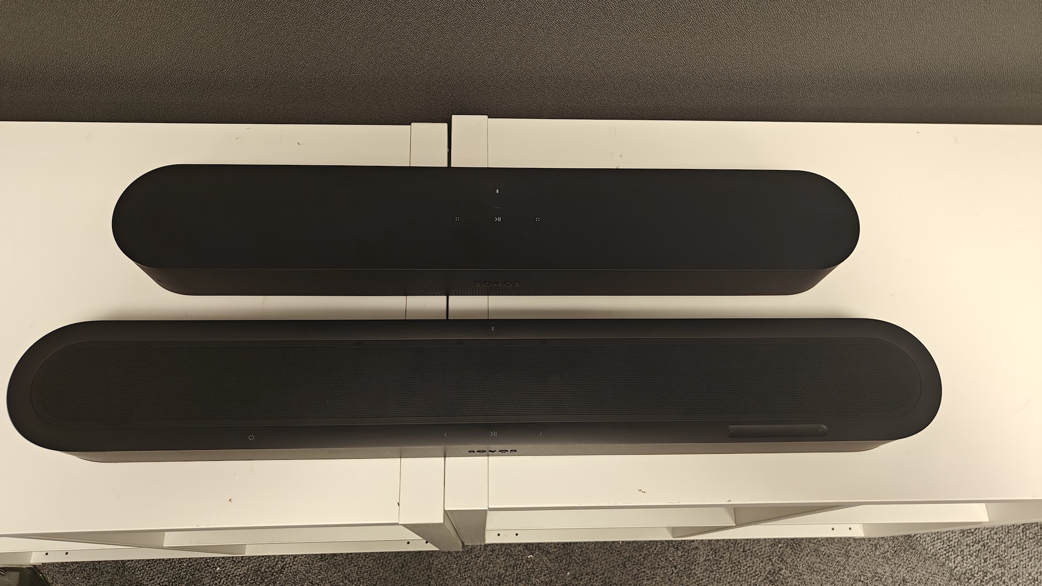 A black Sonos Beam Ultra and Beam (Gen 2) next to each other on a white TV cabinet, shot from directly above.