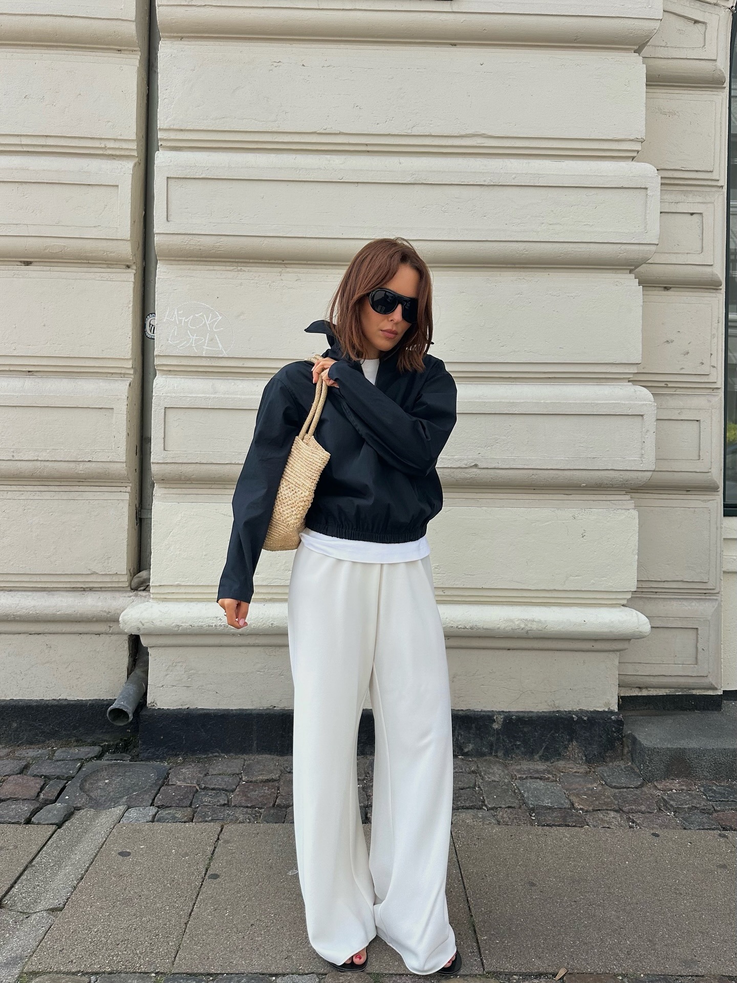 Debora rosa provides her influencer autumn wish list. She wears a white satin trouser, black windbreaker and white tshirt. She carries a raffia bag under her shoulder in a recent image from her Instagram.