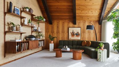 A living room with wood walls, exposed wood beams on the ceiling, a floating shelving system with a sideboard, a curved dark gray couch, a wooden coffee table, a set of stone plinths, potted houseplants, and a floor lamp