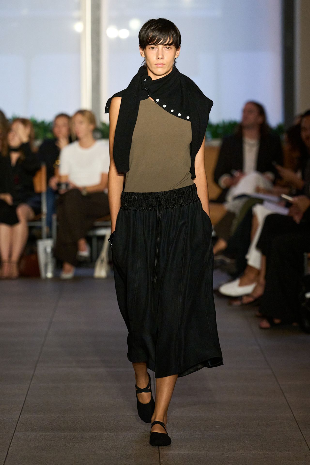 Layering staples for spring 2026 is shown in a photo from Maria McManus's runway show, with a model walking while wearing a black cardigan tied around the shoulders, styled with an olive green tank top, a black midi skirt, and black Mary Jane ballet flats