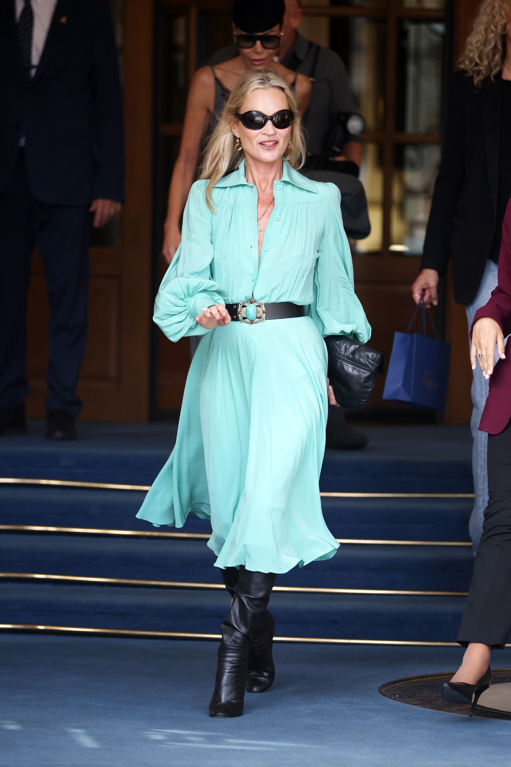 Kate Moss steps outside the Ritz Hotel in Paris during Paris Fashion Week. She wears a turquoise dress with black knee-high boots and a black leather belt.