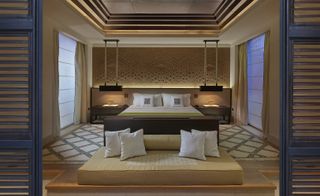 A hotel bedroom with a bed, wooden side tables, a patterned headboard, a floor couch, square stone tiles and multi level square ceilings.