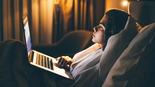 Woman lying in bed on her laptop with bedside light on, finding out how to get back to sleep in the middle of the night