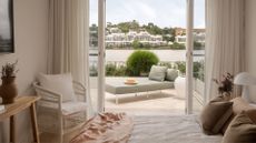 A bright, summery bedroom with a bed in a light bedscape, a corner chair, and a side table with a vase of dried flowers alongside a balcony featuring a sun lounger.