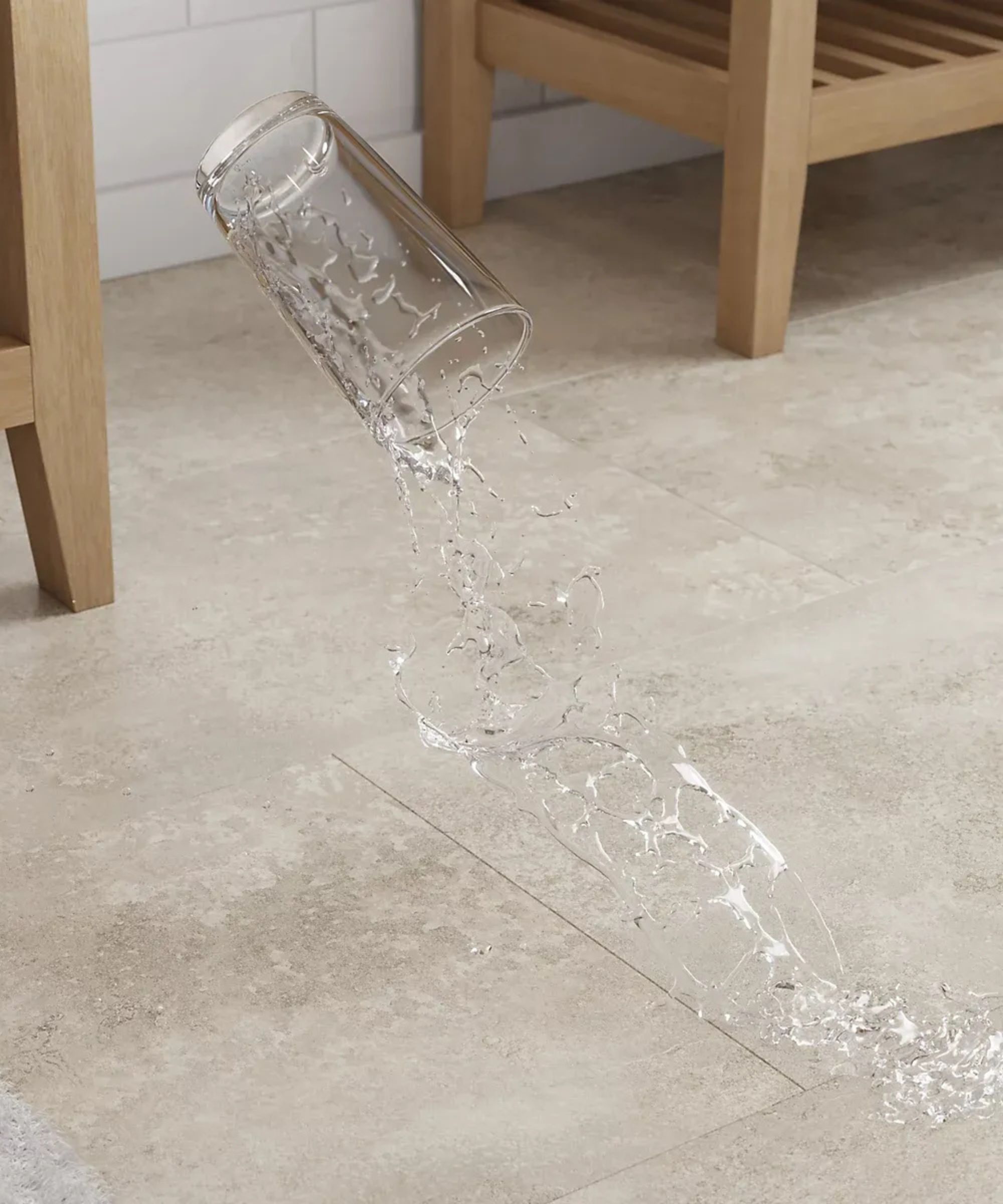 A glass tipping mid-air and spilling water onto a pale stone-effect vinyl floor tile, with wooden furniture legs and white metro tiles visible in the background.