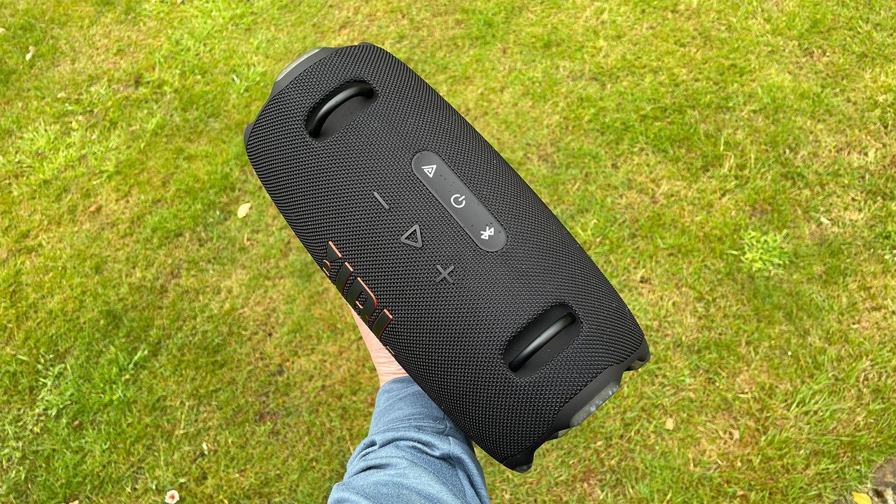 A black JBL Xtreme 4 wireless speaker being held in a hand. A grassy lawn can be seen in the background.