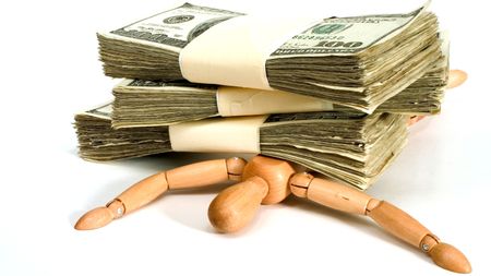 A wooden figure is held down by stacks of hundred-dollar bills.