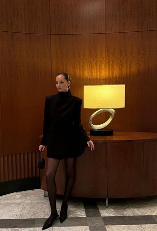 hanukkah outfit idea is shown in a photo of a woman standing inside a lobby wearing a black blazer dress with gold earrings, a black mini bag, black sheer tights, and black pointed kitten heels