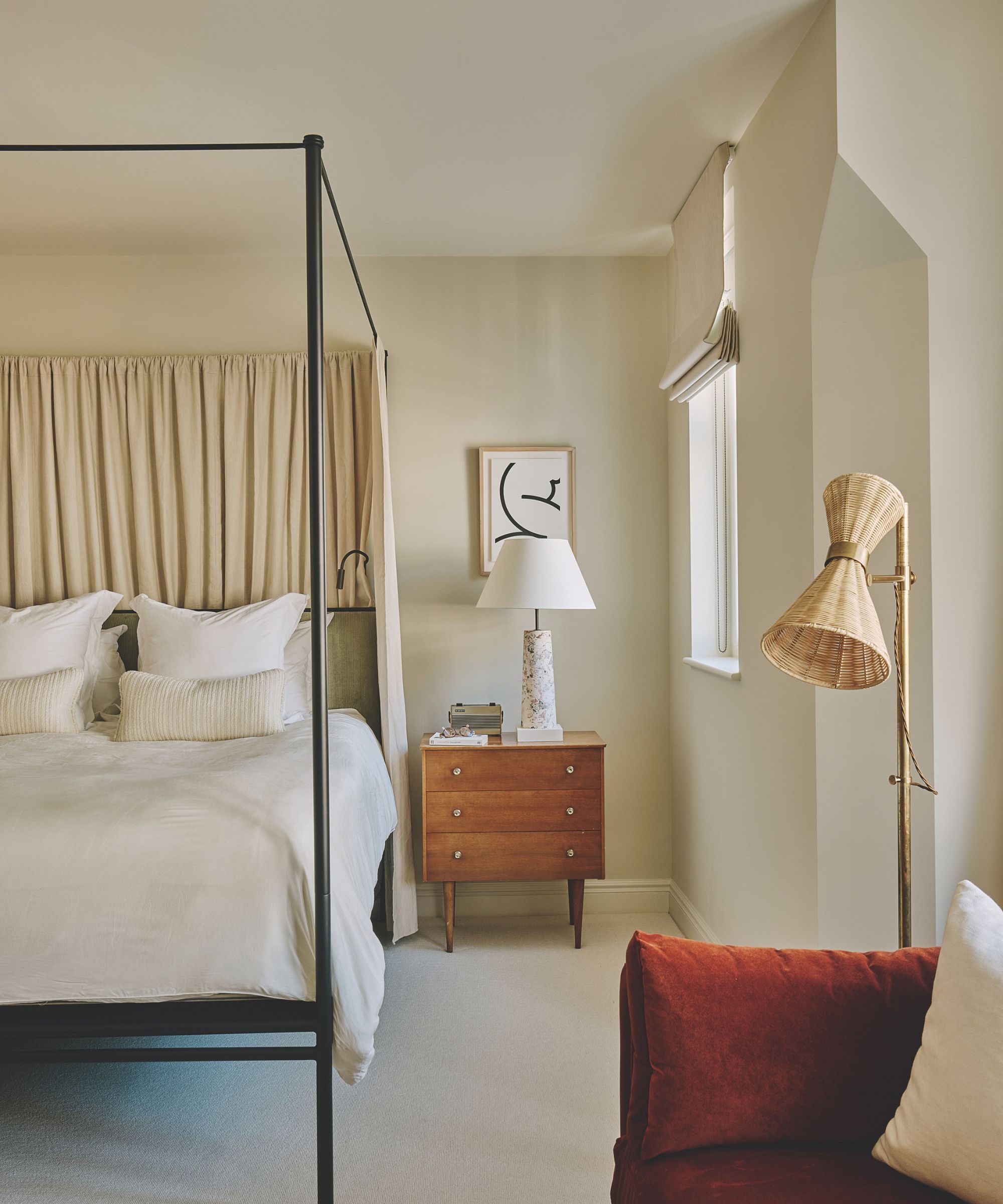 Bedroom with black metal canopy bed, beige fabric headboard, wooden nightstand with table lamp, woven floor lamp, and a red upholstered chair