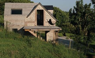 Brick house, windows and doorway, side steps, wooden platform balcony with black chair, hillside, patio area, trees, blue sky