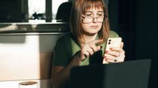 Woman sitting in front of laptop with a cup of coffee next to her is looking at a smartphone