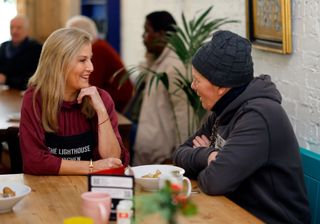 Duchess Sophie wearing a red top and black apron sitting at a table with a woman wearing a black hat and top and smiling