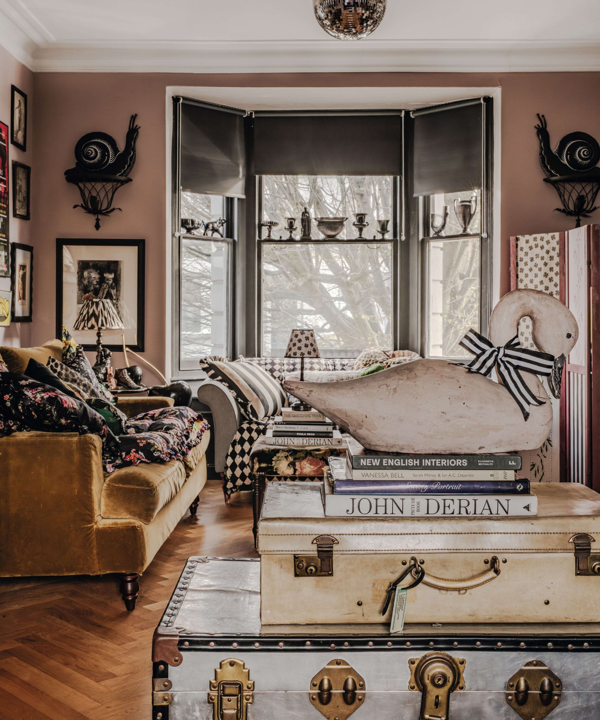 a dusty pink painted living room with bay window with black blinds, ochre velvet sofa and vintage collections