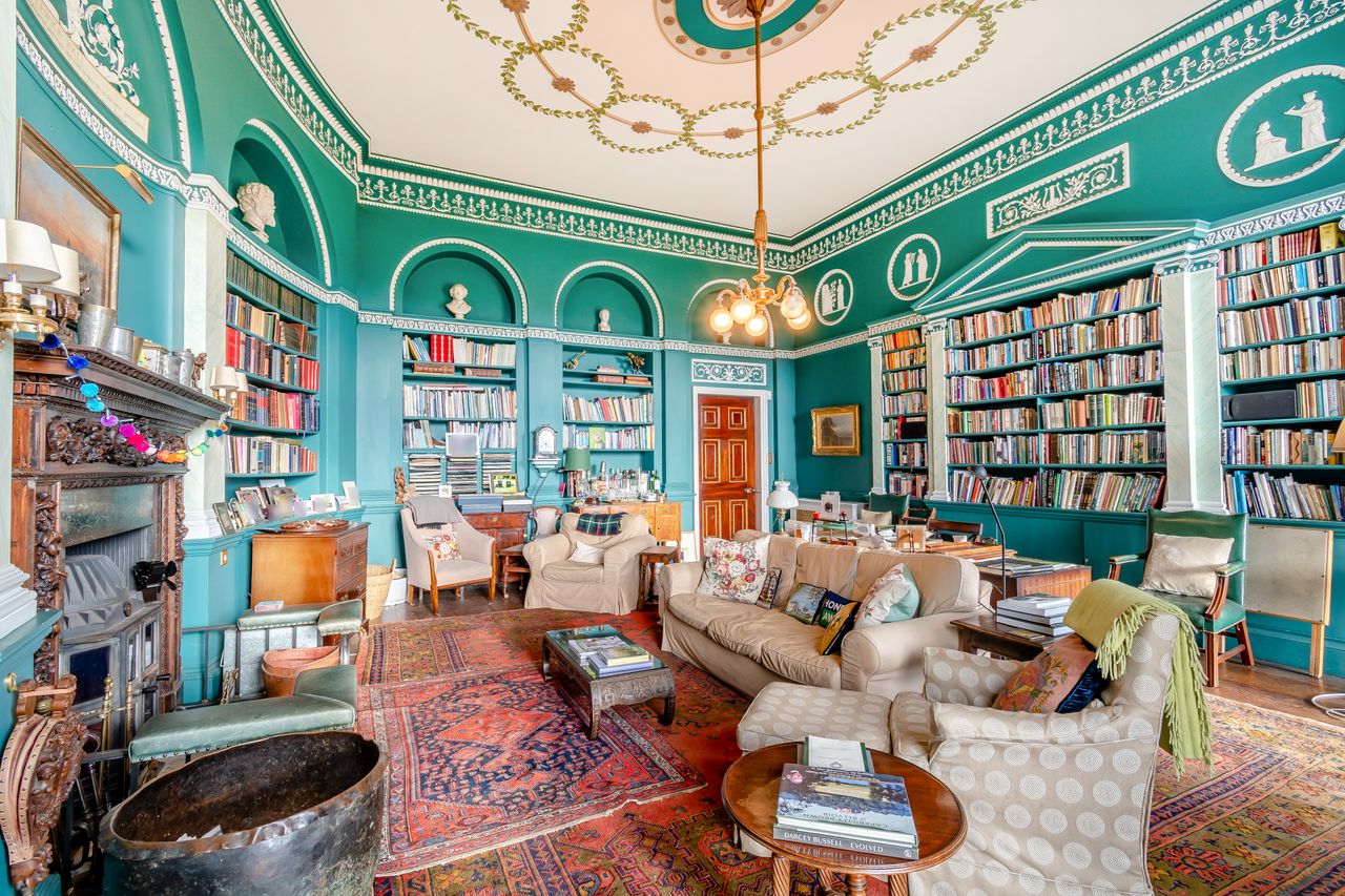 A teal library with an ornate ceiling and many colourful books and pieces of furniture scattered about