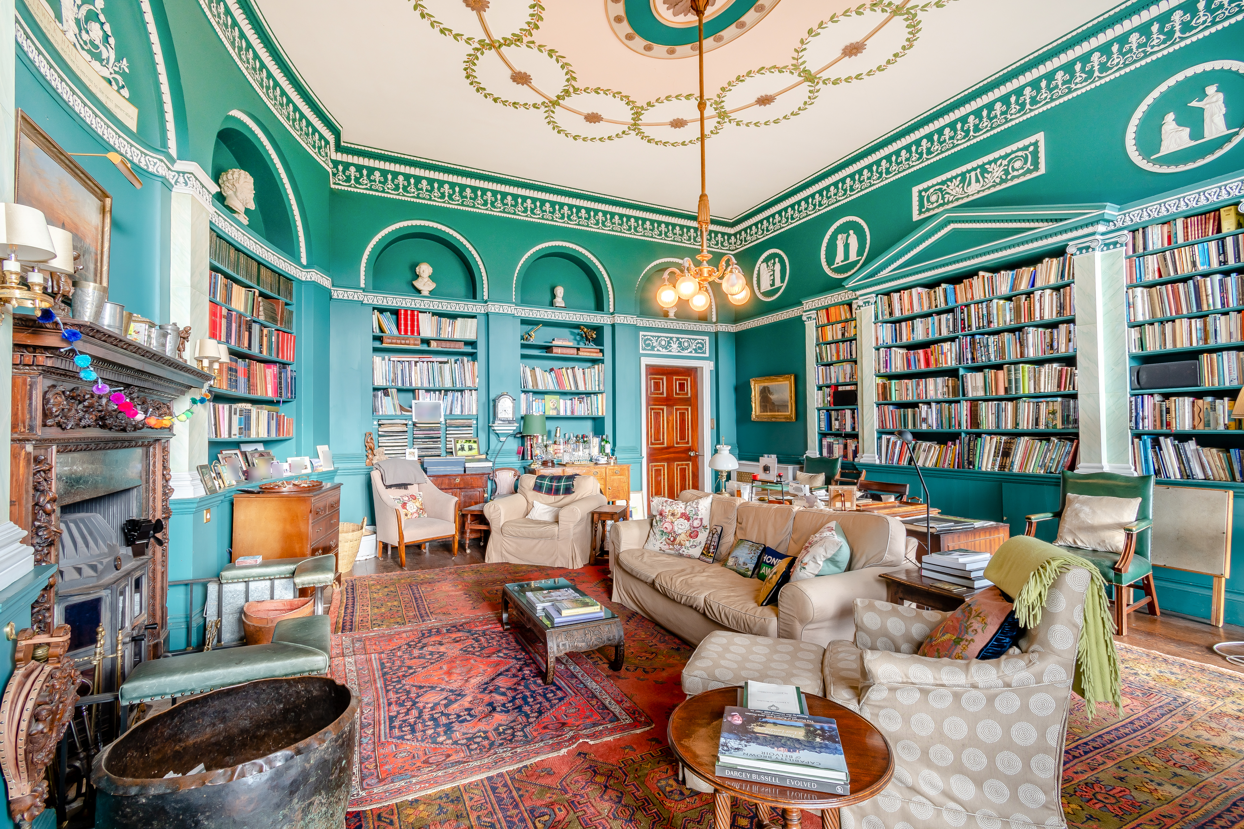 A teal library with an ornate ceiling and many colourful books and pieces of furniture scattered about