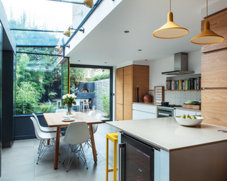 A kitchen extension with big windows with black frames in front of a wooden dining table with white chairs