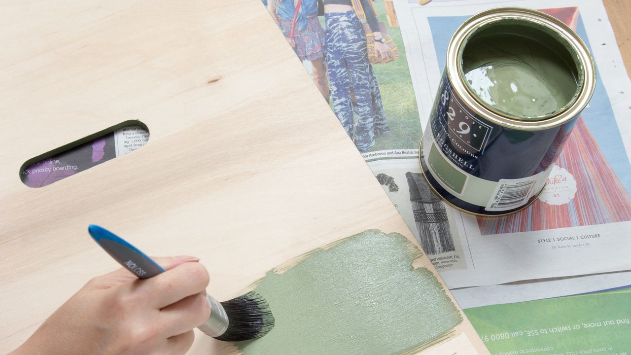 A piece of wood is being painted with green paint with an open tin next to it placed on top of newspaper