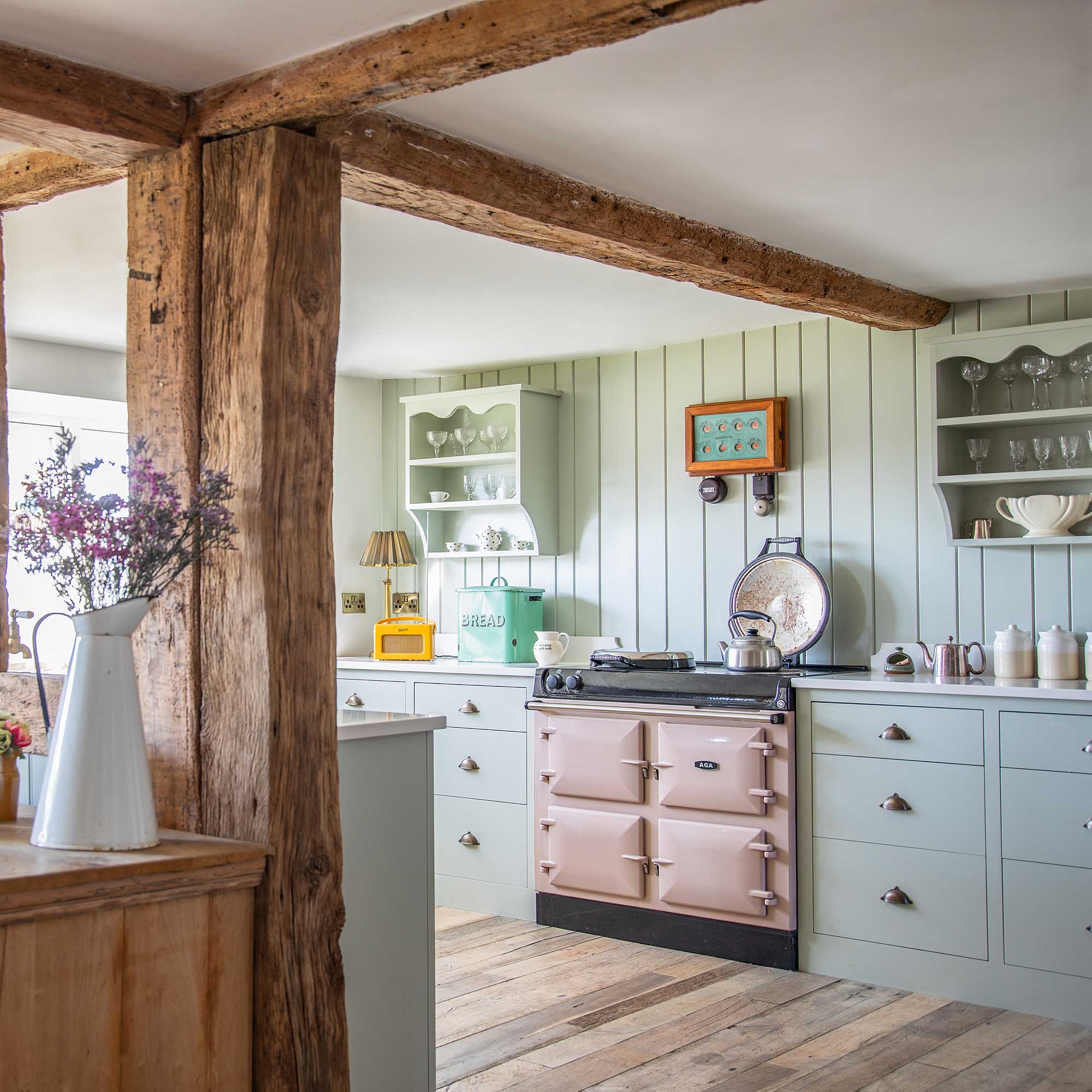country kitchen with tongue and groove splashback, pink range cooker, green cabinets, wooden beams