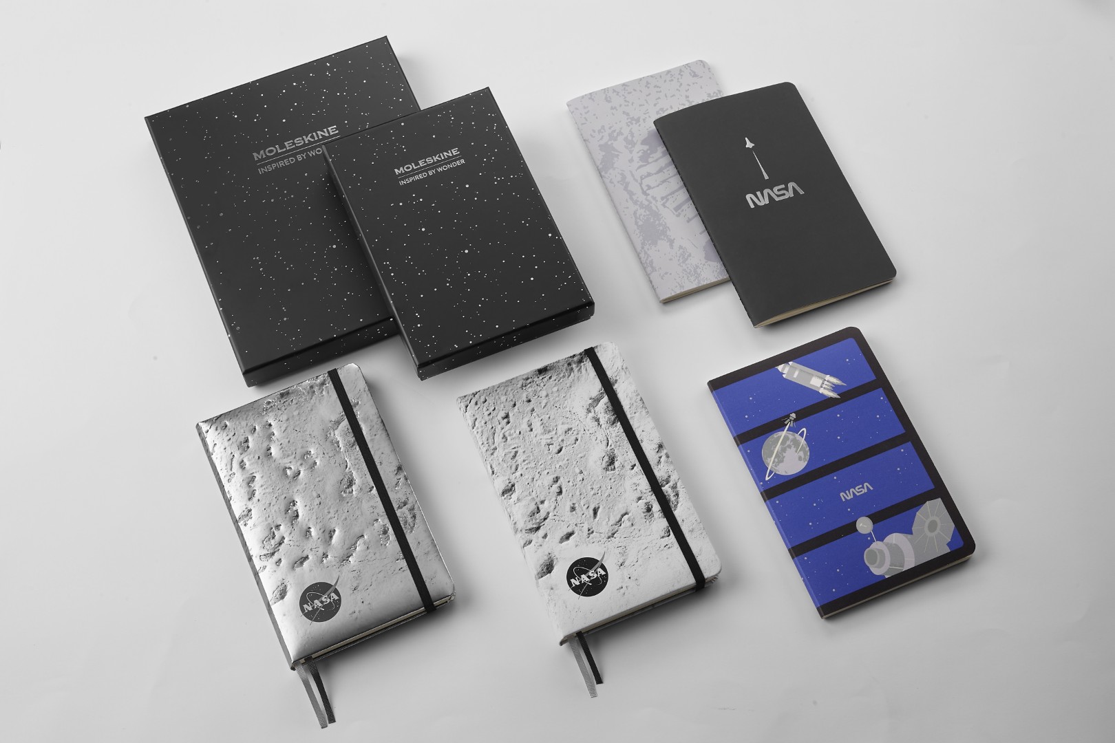 Full collection of NASA-inspired Limited Edition Notebooks from Moleskine: Black gift box with silver stars, 2 softcover notebooks (one with Neil Armstrong's moon footprint and the other is black with a minimalist rocket blasting up and "NASA" text, 2 hardcover notebooks showing off the moon's surface (silver and dark silver), and the softcover, glow-in-the-dark Volant journal.